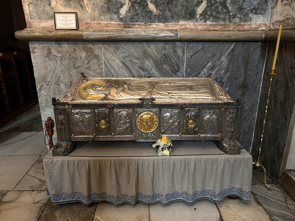 A silver embellished sarcophagus with intricate carvings and a golden circular emblem, placed on a draped table in a stone interior, with a candle holder and a small bouquet of flowers nearby.