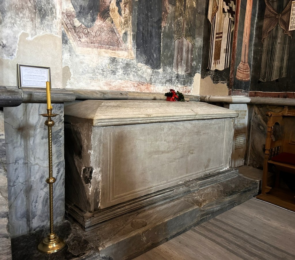 An ancient stone tomb with a simple design, resting on a marble platform, accompanied by a candle holder and a small bouquet of flowers, set against a wall adorned with faded religious frescoes.