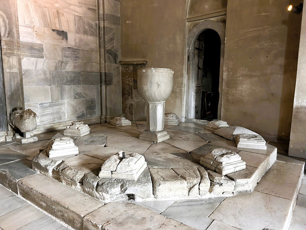 An ancient stone basin in a marble-lined room, surrounded by stone remains and structures, with an arched doorway in the background.