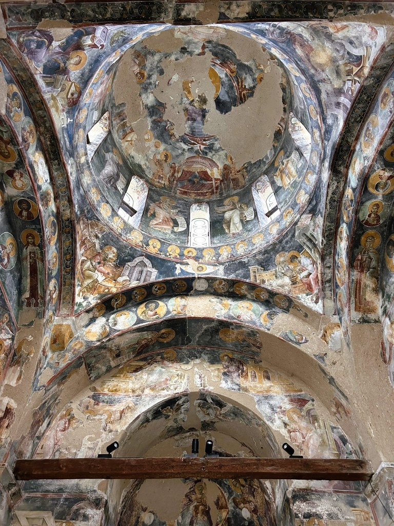Ceiling of a historic church featuring ancient frescoes, with religious figures and decorative elements, showcasing wear and partial damage.