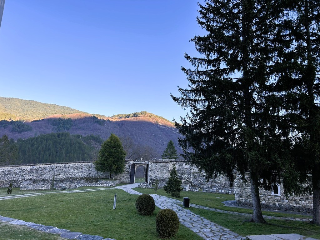 A picturesque scene featuring a stone wall and gate surrounded by neatly maintained green grass, trees, and distant hills under a clear blue sky.