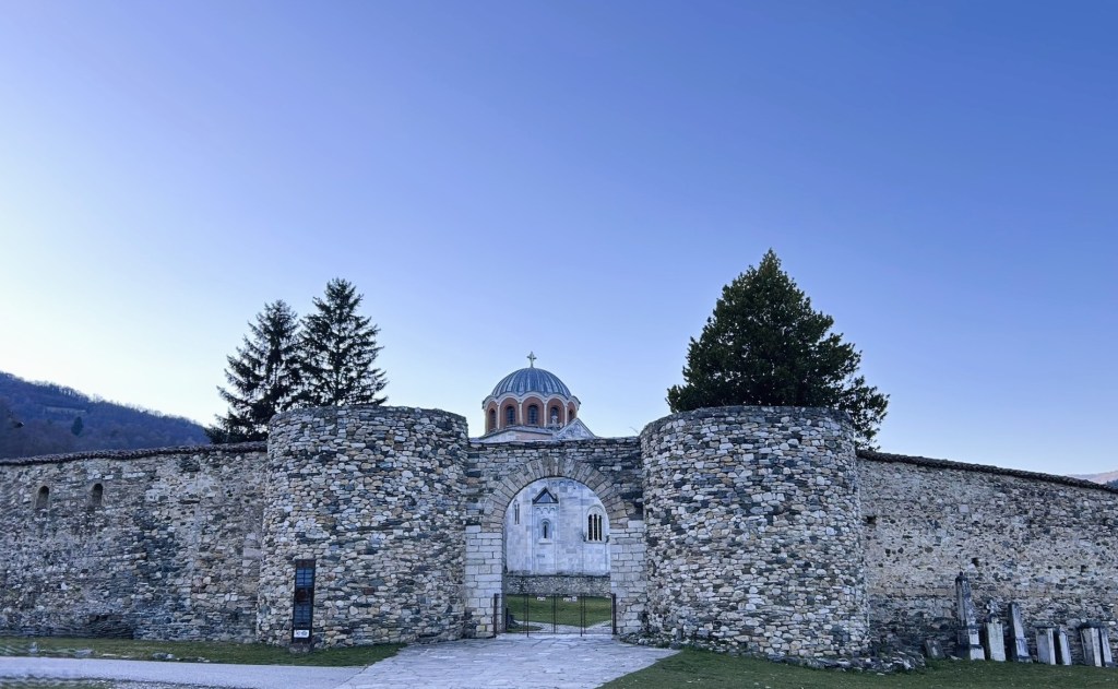 A historic stone fortress with a domed church visible in the background, surrounded by tall trees and a clear blue sky.