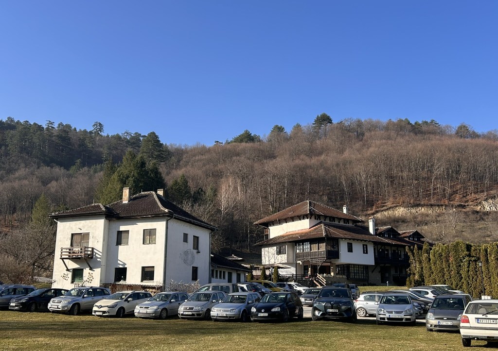 A scenic view of two traditional buildings surrounded by a parking lot filled with parked cars, set against a backdrop of a forested hillside under a clear blue sky.