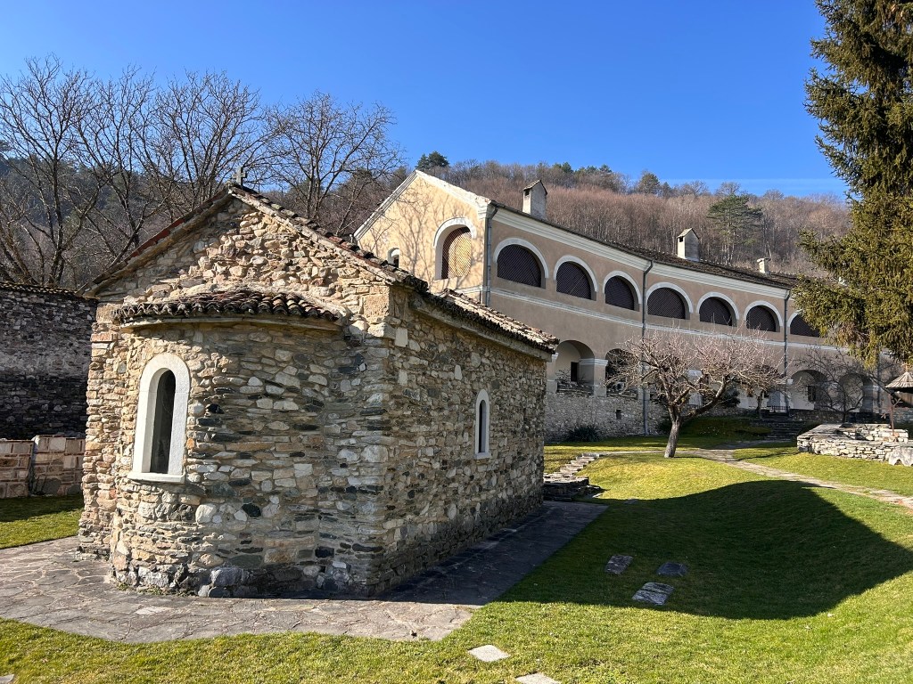A stone chapel with a curved roof and arched windows, surrounded by green grass and trees, with a larger building in the background under a clear blue sky.