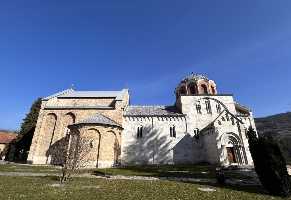 Historic stone church set against a clear blue sky, featuring a distinct dome and arched doorways, surrounded by green grass and trees.