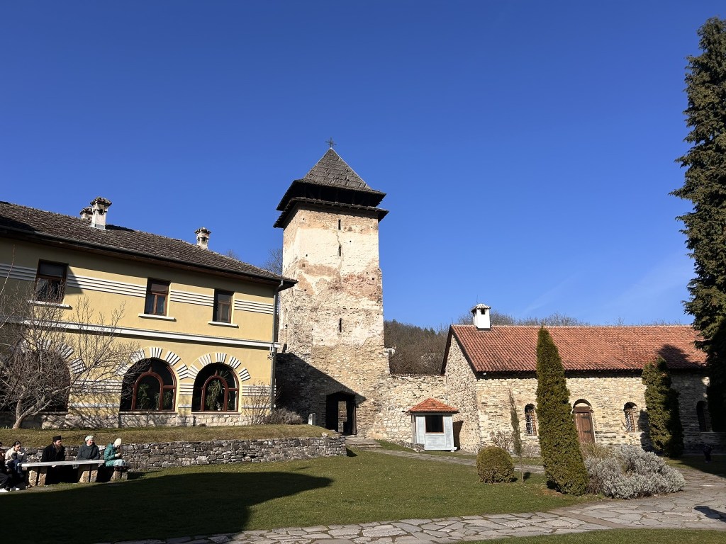 Historic buildings with a stone tower and adjacent structures under a blue sky, surrounded by greenery and visitors sitting on a bench.