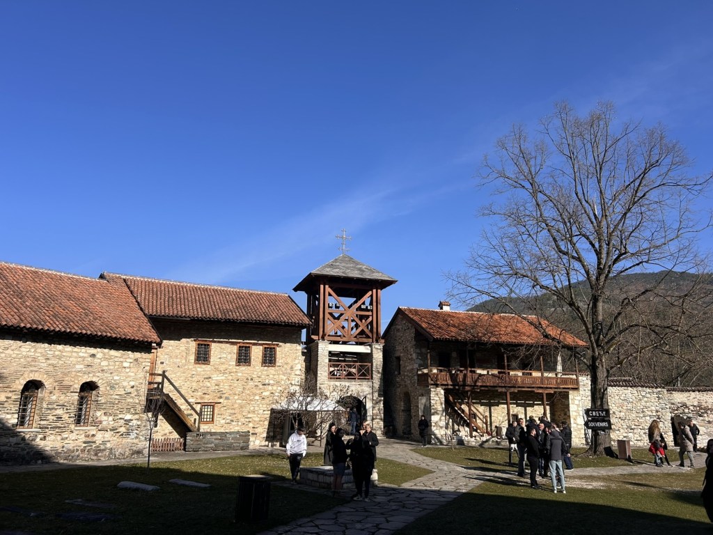 View of a historic stone building complex with a wooden bell tower, surrounded by visitors and bare trees against a clear blue sky.