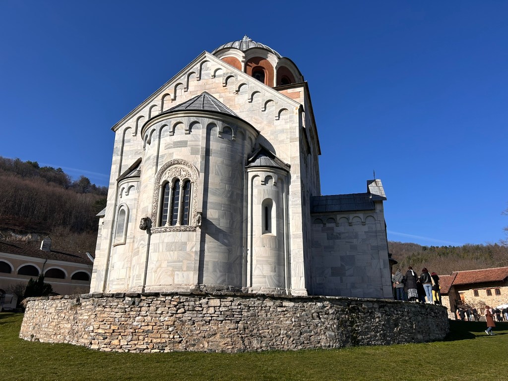 A historic stone church with decorative windows and a round tower, set against a clear blue sky, surrounded by visitors and greenery.