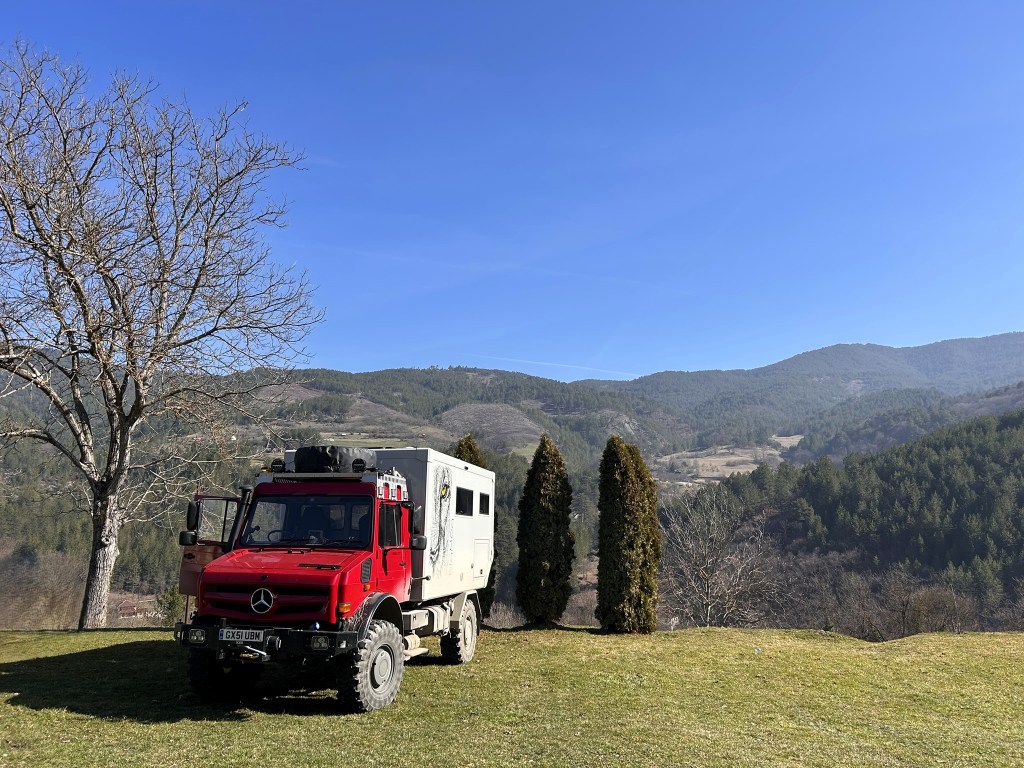 A red Mercedes off-road vehicle parked on green grass with mountains in the background and a clear blue sky.