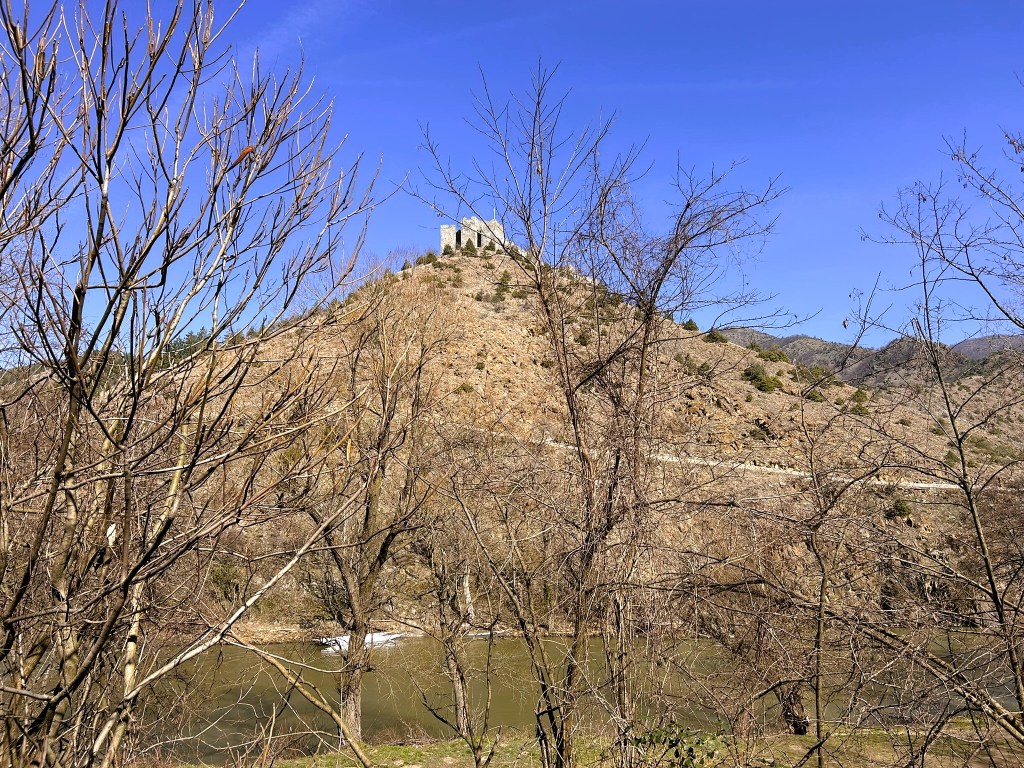 A hill with a castle ruins at the top, surrounded by bare trees and a body of water in the foreground, against a clear blue sky.