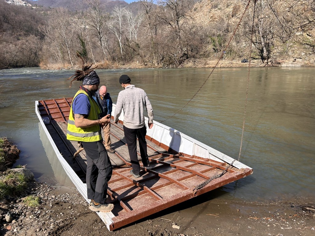 Three men boarding a flat-bottom boat on a river, surrounded by trees and hills.