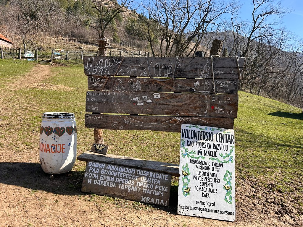 A wooden information board with maps and texts in Serbian, located in a green outdoor area. The board highlights various local attractions and activities. Beside it, there is a painted barrel labelled for donations.