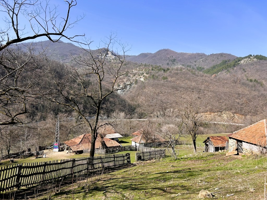 A scenic view of a rural landscape featuring several rustic homes with red roofs, surrounded by bare trees and distant mountains under a clear blue sky.
