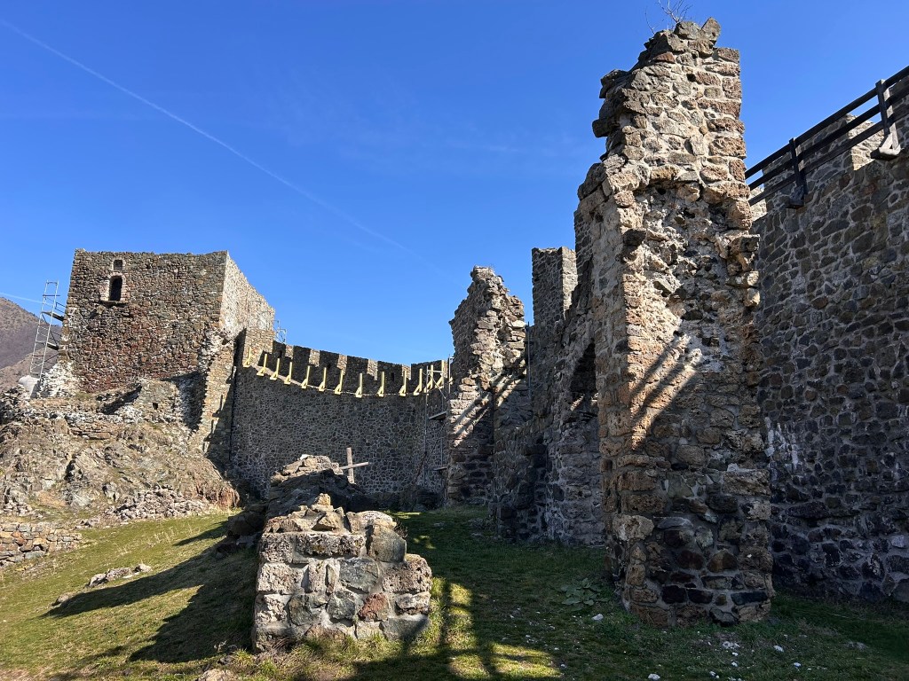 Ruins of a stone castle under a clear blue sky, with partially collapsed walls and grassy areas.