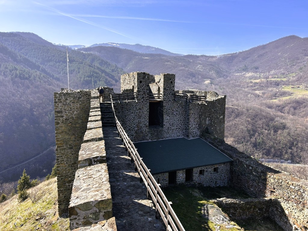 Aerial view of a medieval stone castle ruin perched on a hilltop, surrounded by lush mountains and a clear blue sky.