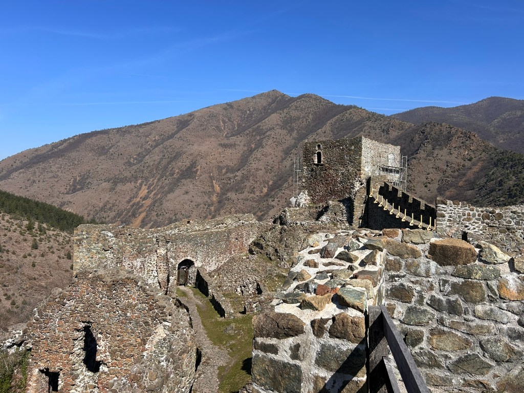 A view of ancient stone ruins atop a hill with mountains in the background under a clear blue sky.