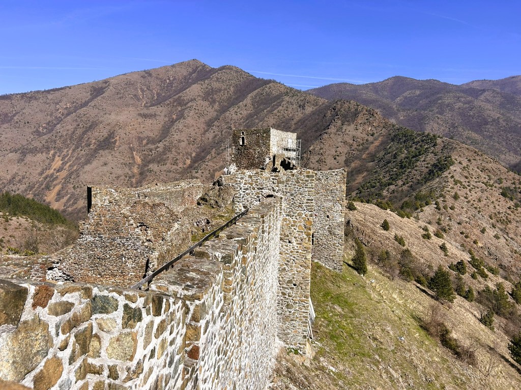A panoramic view of an ancient stone fortress perched on a hill, surrounded by rolling mountains and a clear blue sky.