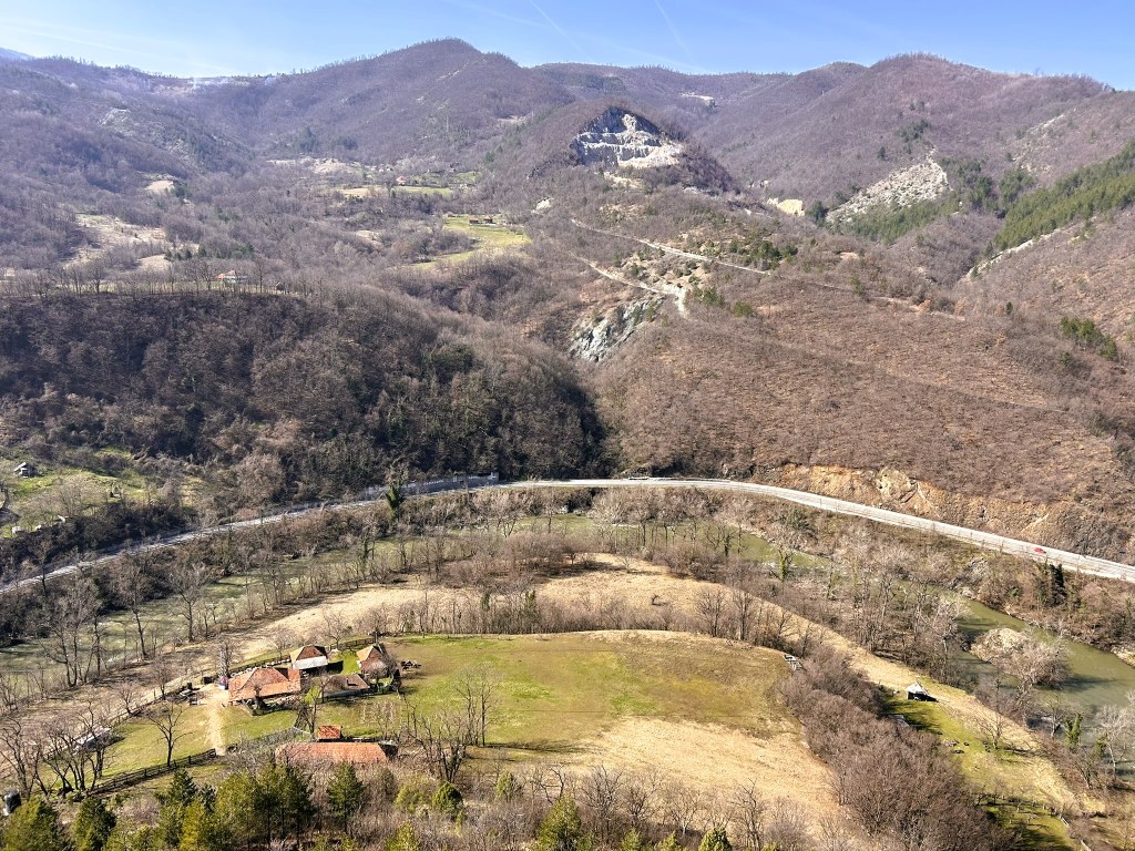Aerial view of a rural landscape featuring a grassy field, a small cluster of houses with red roofs, and a winding road alongside a river, surrounded by bare hills and sparse trees.