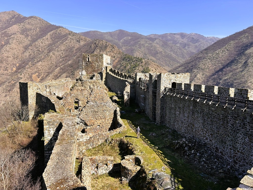 A scenic view of ancient stone castle ruins with a mountainous landscape in the background under a clear blue sky.