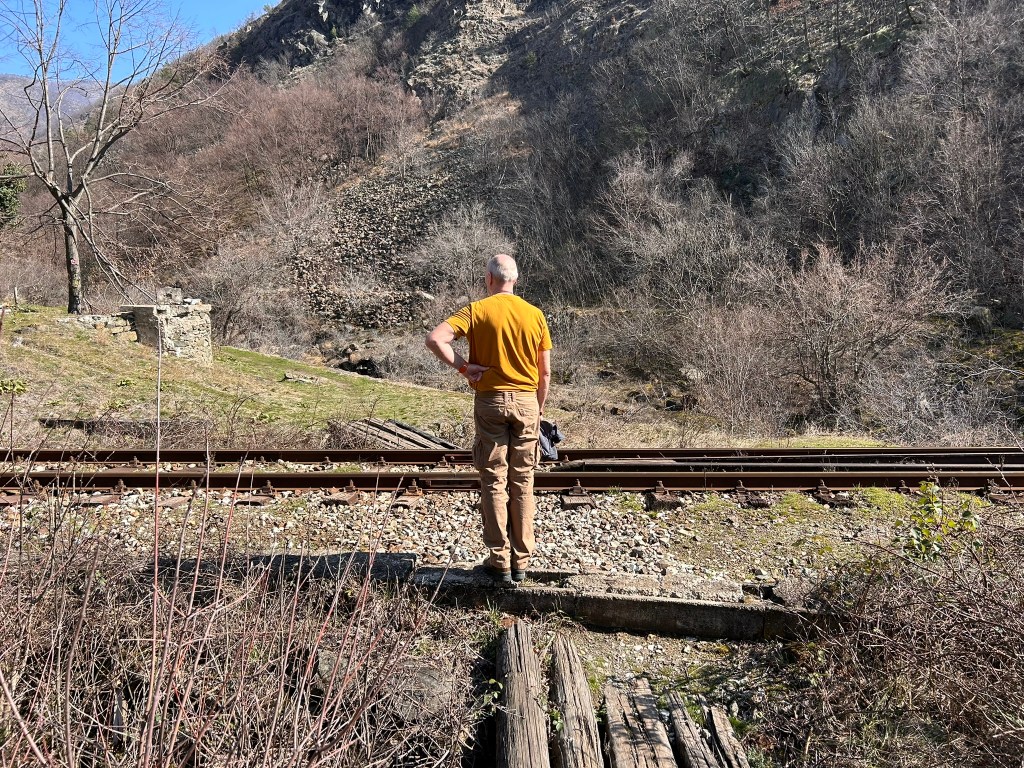 A person standing on a railway track, facing a mountainous landscape with sparse vegetation and trees. The individual is wearing a yellow shirt and has their hands on their hips, contemplating the scenery.