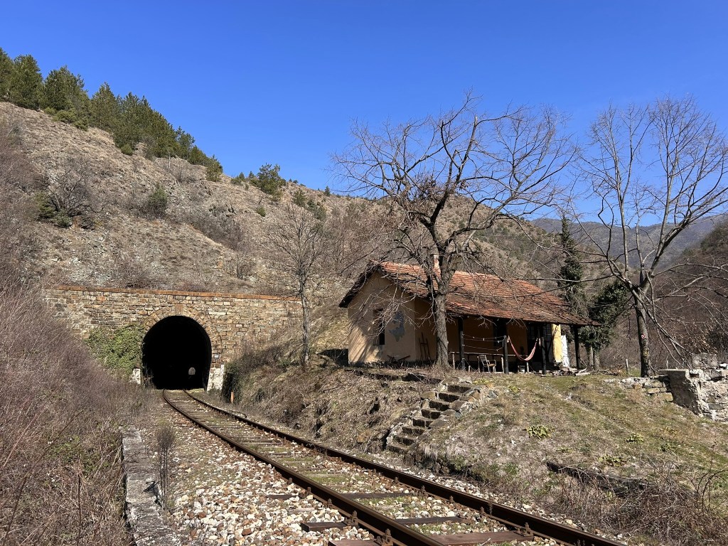 A scenic view of an old stone tunnel at the foot of a hill, with a small abandoned house nearby and railway tracks leading towards the tunnel. The landscape is semi-barren with sparse trees and a clear blue sky.