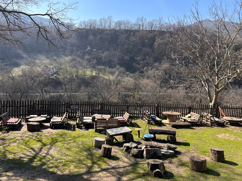 A scenic outdoor seating area with wooden benches and tables, surrounded by nature and a wooden fence, overlooking a hillside with trees and a distant house.