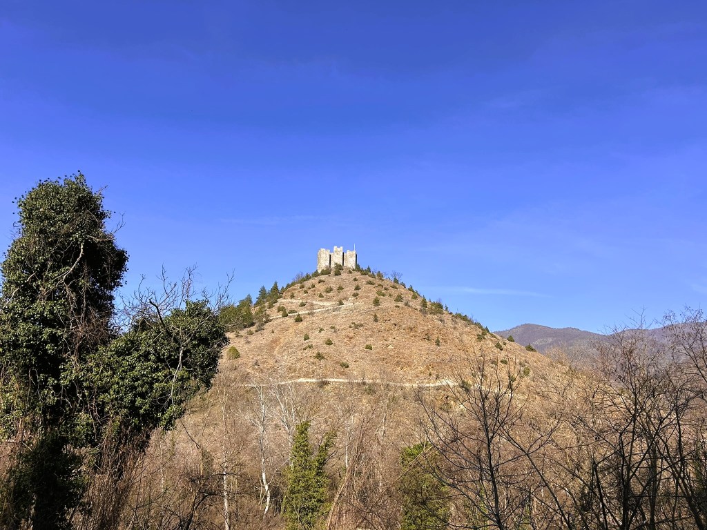 A view of a historic castle situated atop a grassy hill under a clear blue sky, surrounded by sparse trees and rugged landscape.