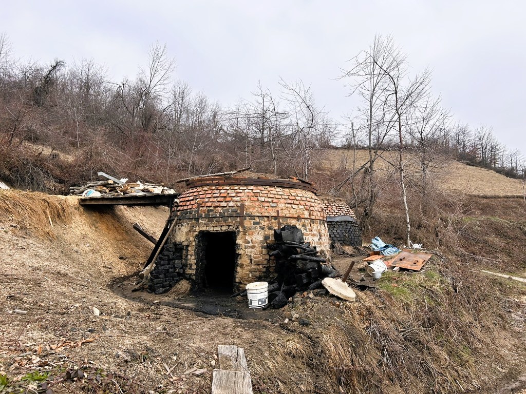 A traditional brick and stone oven structure situated on a hillside, surrounded by sparse trees and dry vegetation.