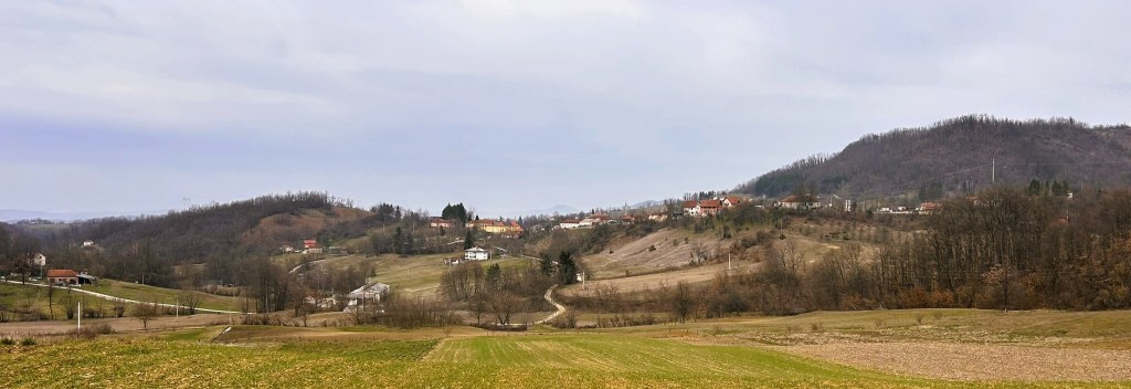 Panoramic view of a rural landscape featuring rolling hills, scattered houses, and fields in various shades of green and brown, under a cloudy sky.