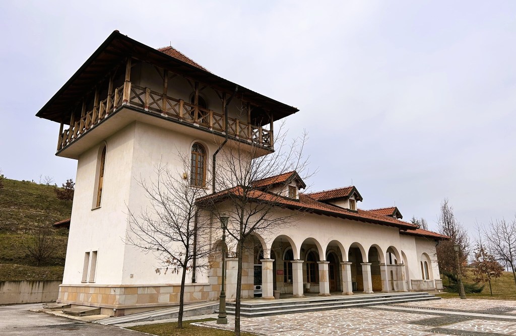 A two-storey building featuring a combination of arched windows and wooden balconies, surrounded by bare trees and a grassy slope.