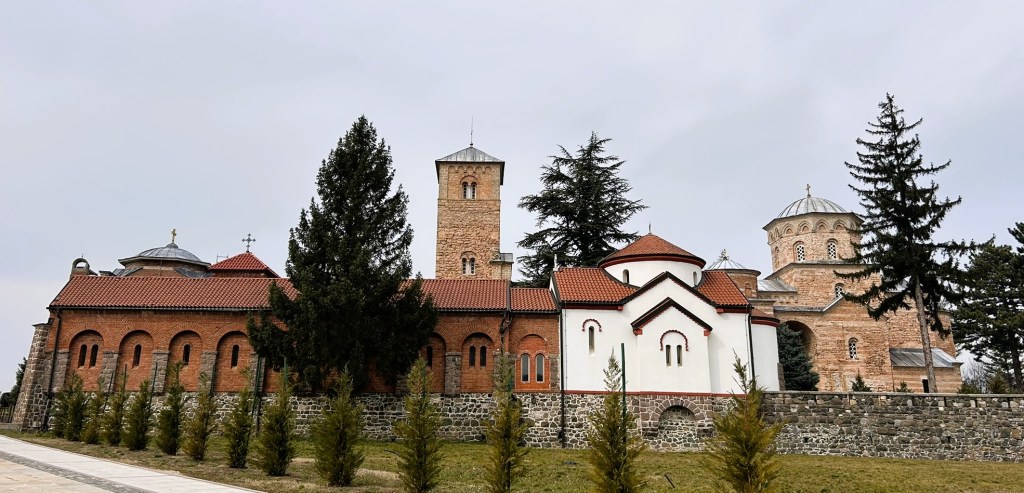 A view of a historical monastery with red brick buildings and a prominent tower, set against a cloudy sky and surrounded by evergreen trees.