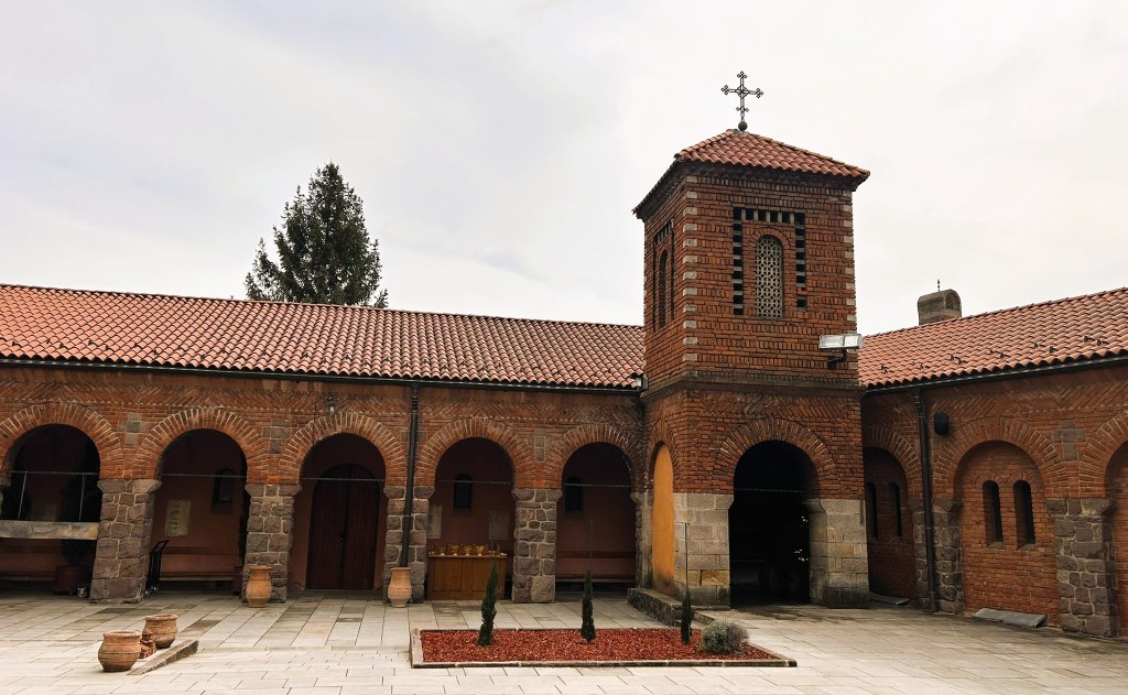 Exterior view of a brick church with a bell tower and a tiled roof, surrounded by arches and small trees in a courtyard.