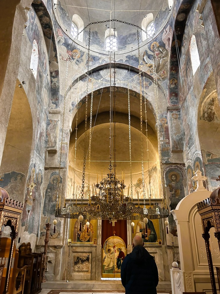 Interior view of an ornate church featuring a high dome, intricate frescoes on the walls, and a prominent chandelier, with a person standing in the foreground.