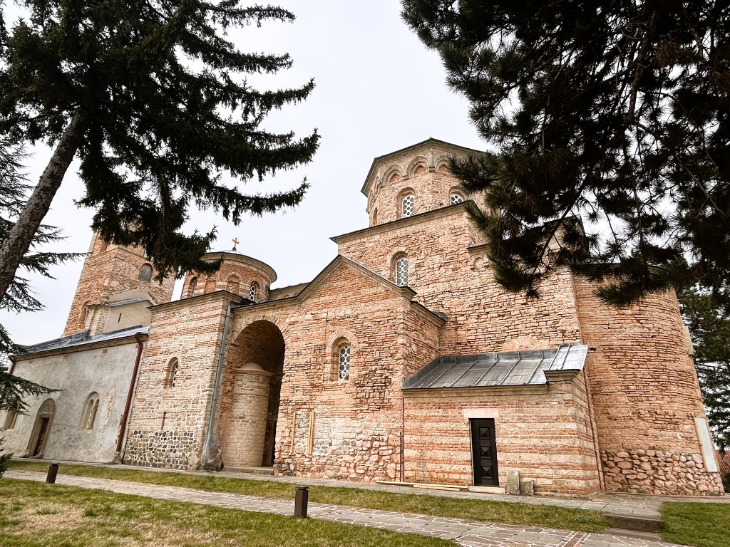 A historic stone church with a large arched entrance and multiple domes, surrounded by tall trees and greenery.