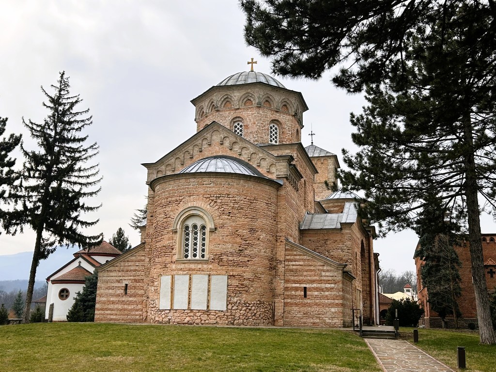 A historic stone church with a dome and cross on top, surrounded by trees and grass, with additional buildings visible in the background.