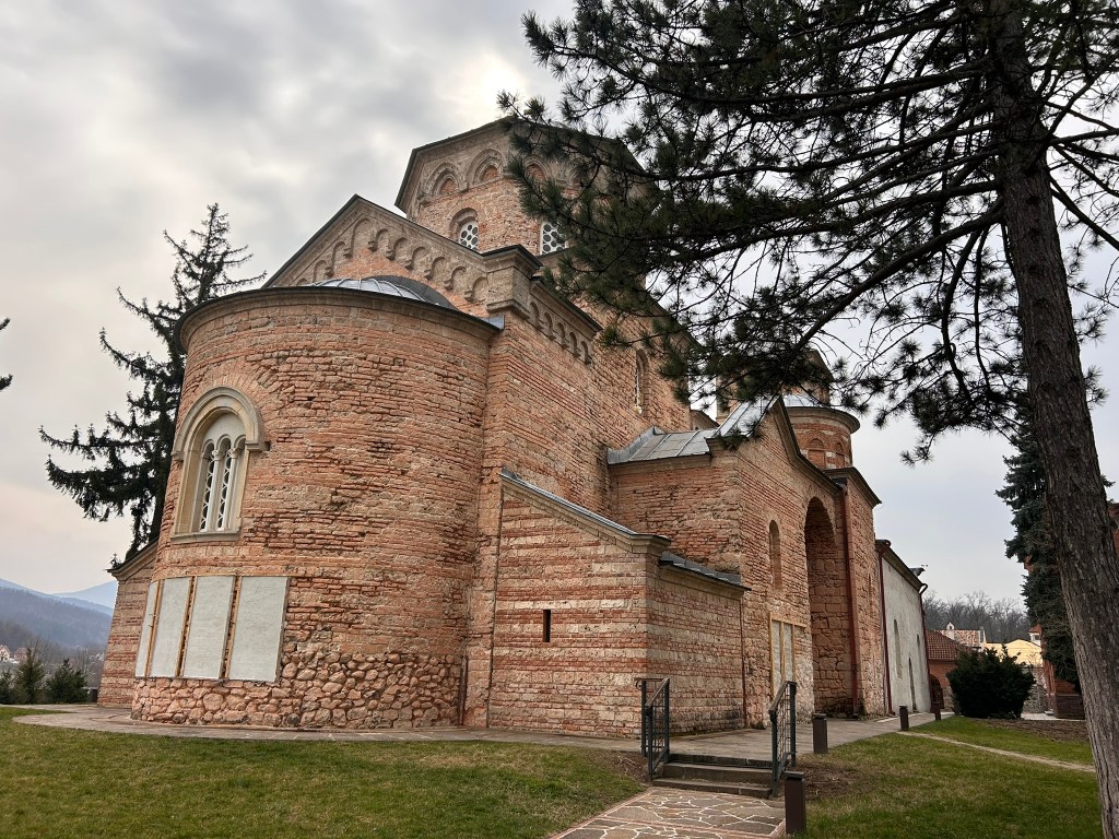 A historic stone church with a rounded architecture, surrounded by trees and grass, under a cloudy sky.