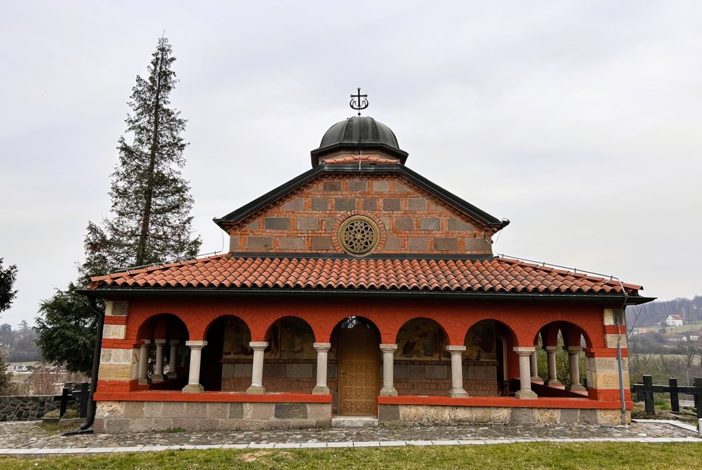 Exterior view of a small church featuring a red and stone façade with a tiled roof, arched entrances, and a central dome topped with a cross.