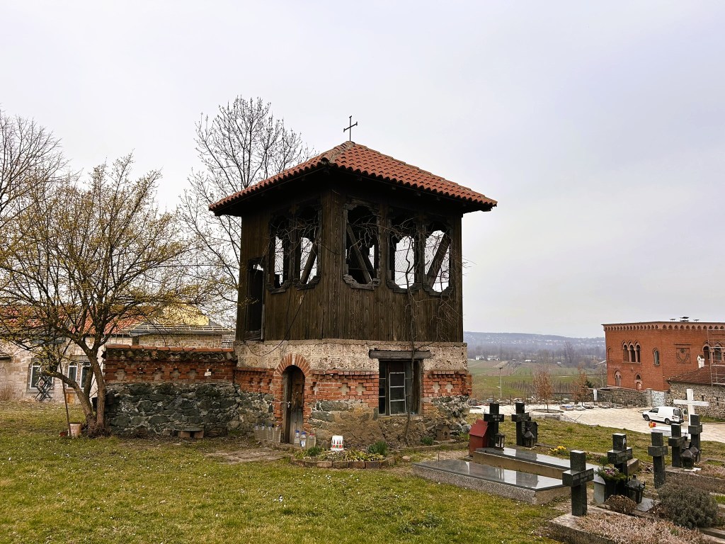 Historic bell tower with a wooden structure and tiled roof, surrounded by trees and a cemetery, under a cloudy sky.