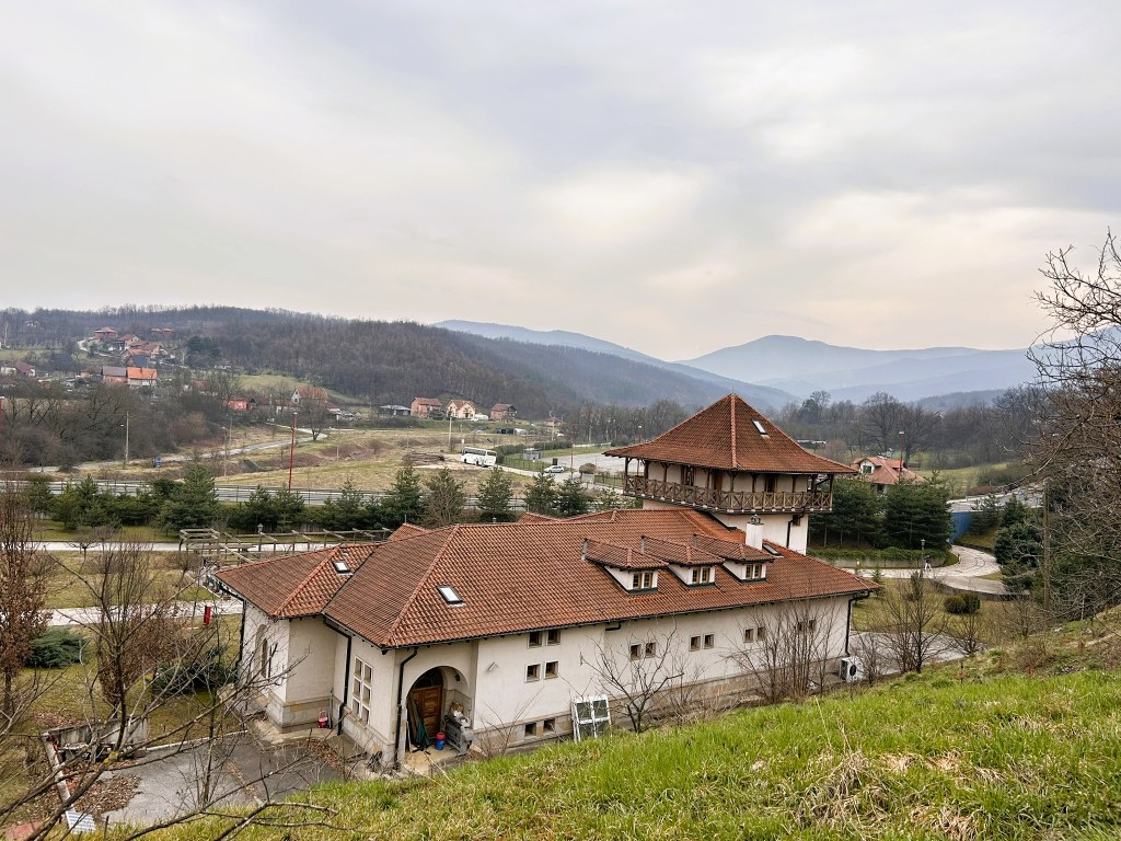 A view of a rustic building with a distinctive roof, surrounded by rolling hills and scattered houses in a rural landscape under an overcast sky.