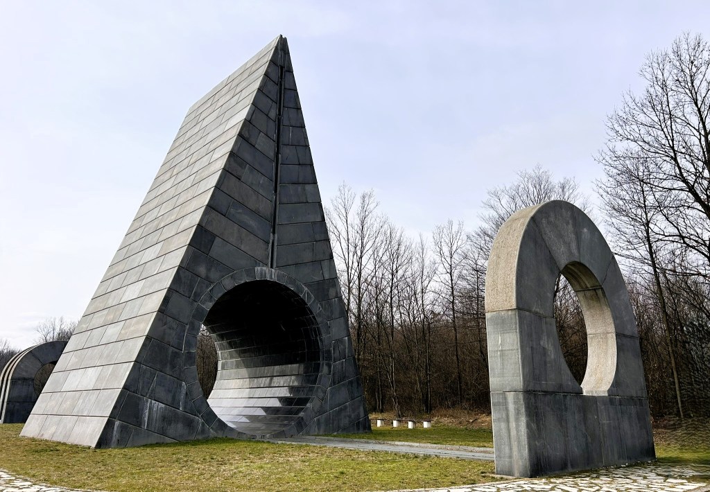 A large, modern stone sculpture featuring an angular pyramid with a circular opening, alongside a circular arch, set in a grassy area with bare trees in the background.