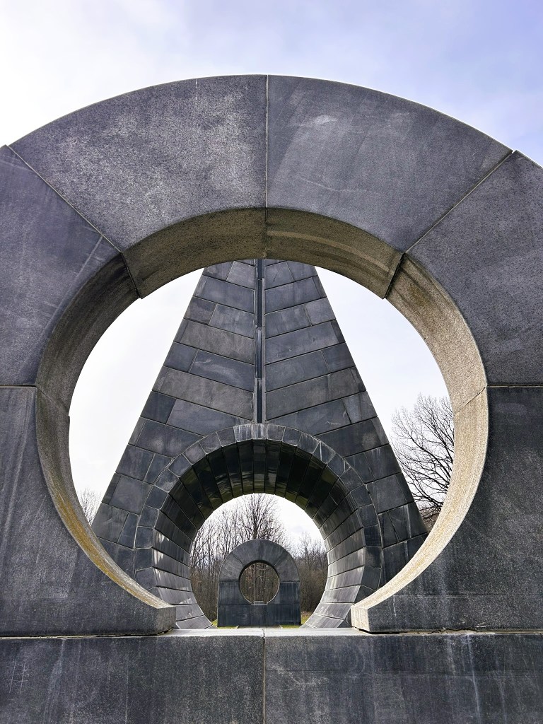 A large, abstract stone sculpture featuring angular and circular shapes, set against a cloudy sky.