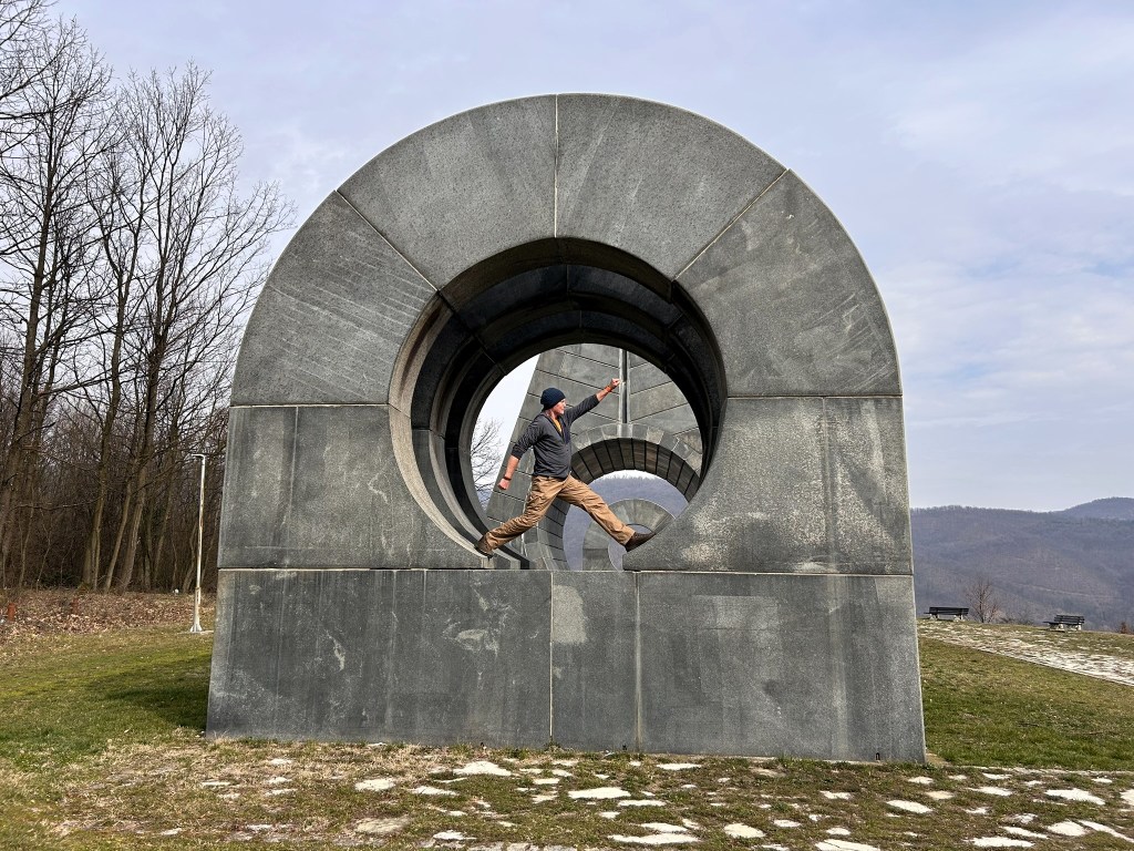 A person leaping through a large circular stone sculpture set in an outdoor landscape with trees in the background.