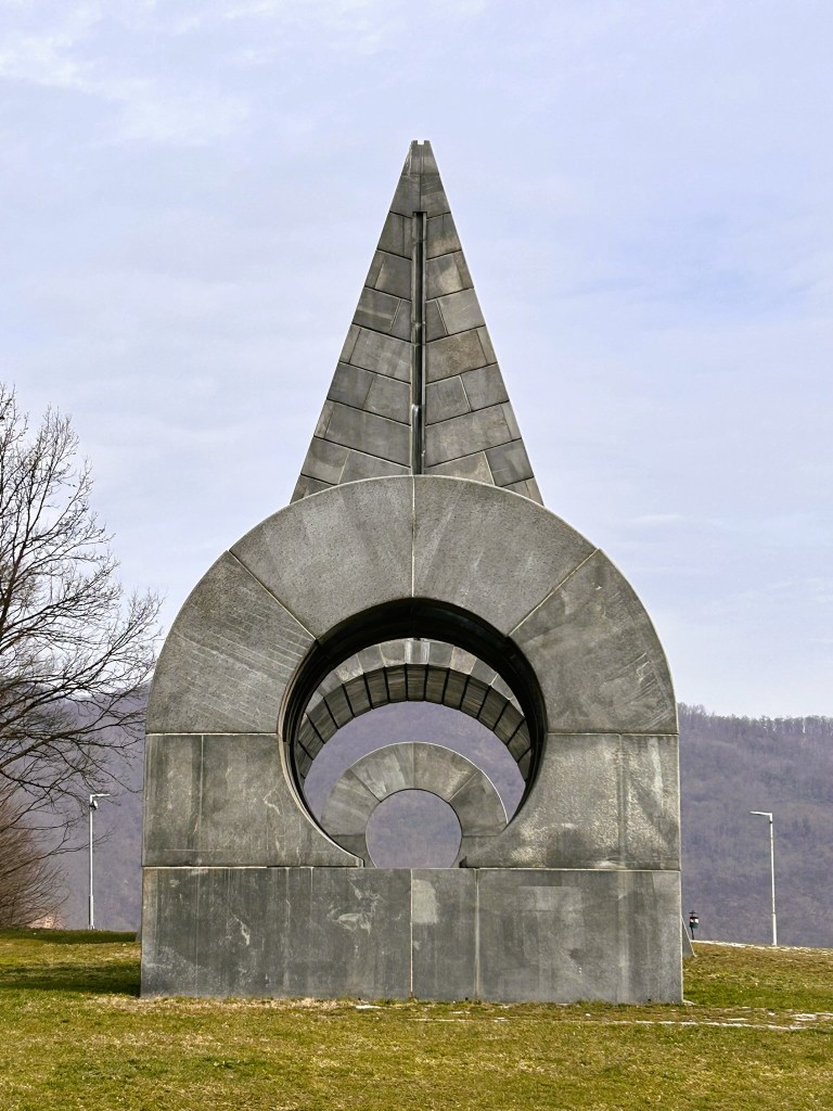A large, abstract concrete sculpture resembling a pointed arch with circular cutouts, set against a cloudy sky and grassy landscape.