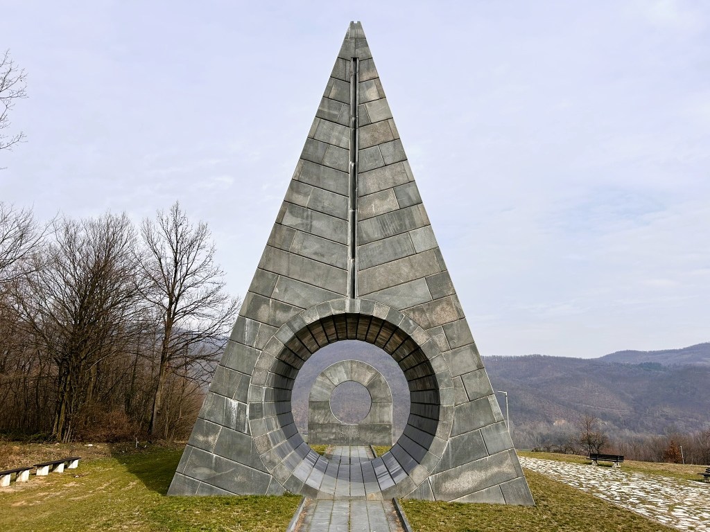 A tall, triangular stone monument with a circular opening at its base, surrounded by trees and hills in the background.