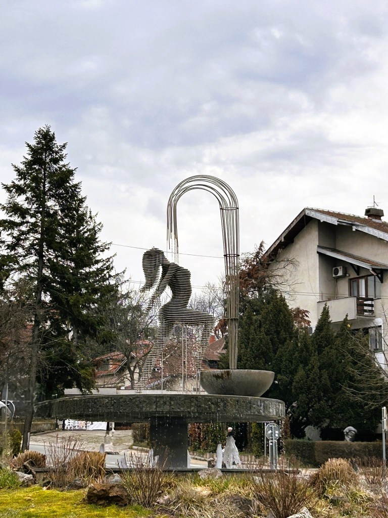 A modern sculpture resembling a figure in motion, made of curved metal rods, set on a circular base with a water feature, surrounded by greenery and residential buildings.