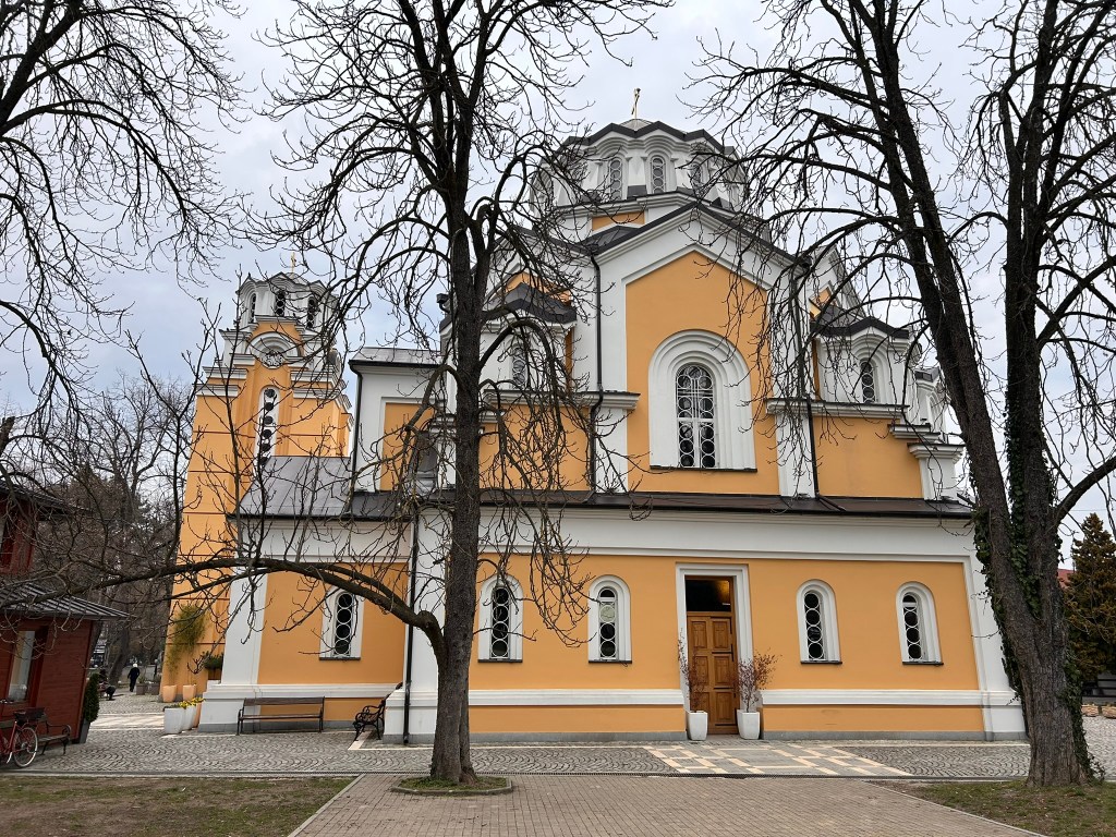 A yellow and white church building with multiple domes, surrounded by leafless trees and a paved pathway.