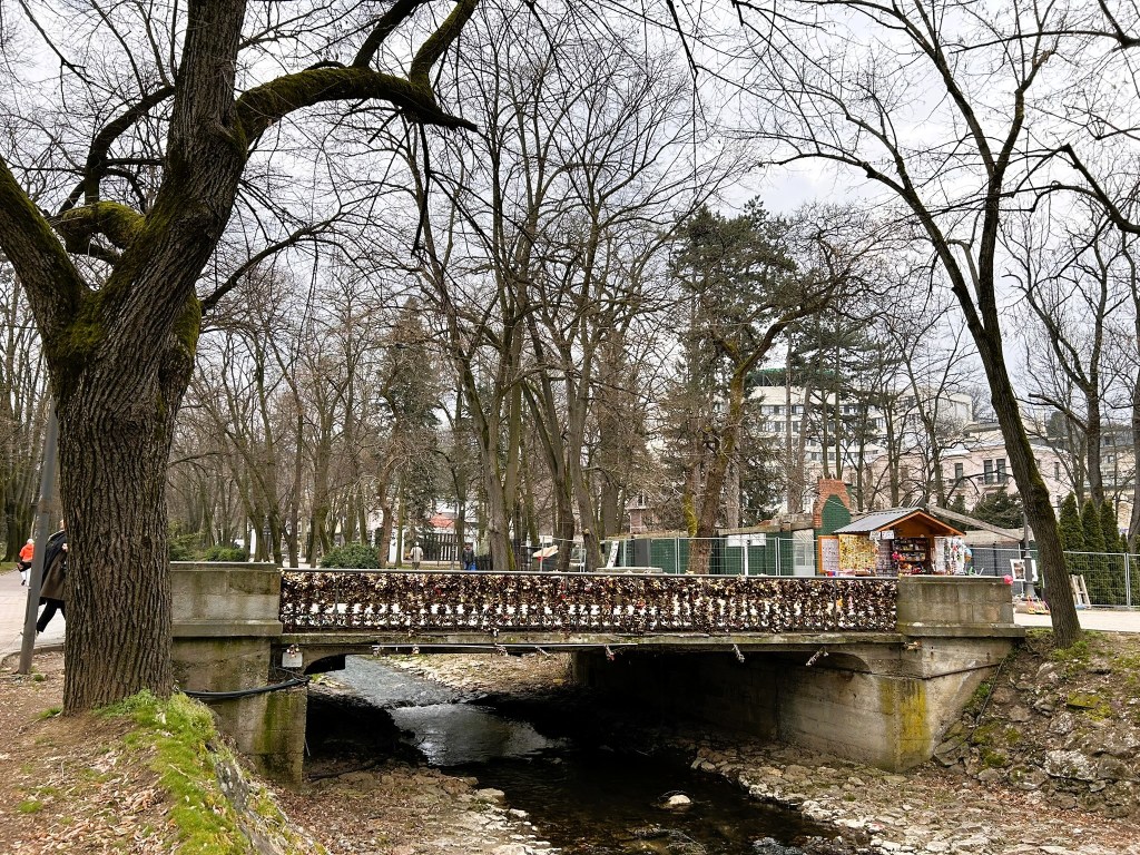A concrete bridge adorned with locks crosses a small stream, surrounded by leafless trees in a park setting.