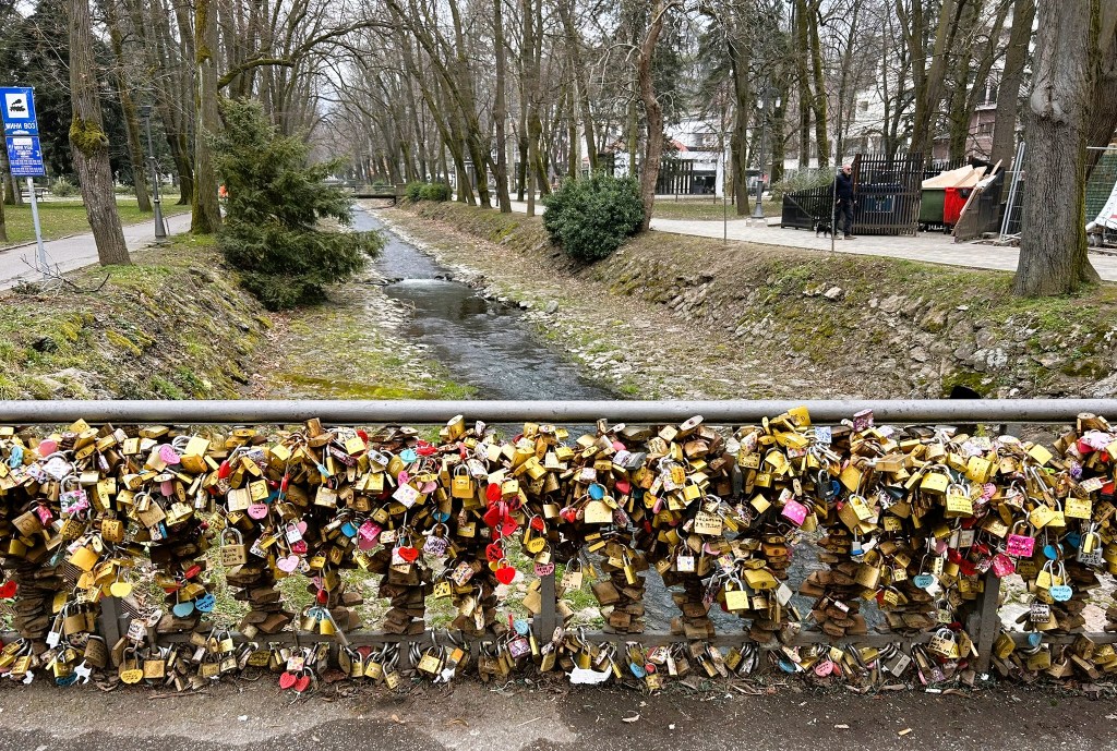 A bridge adorned with numerous love locks, overlooking a small stream lined with trees in a park setting.