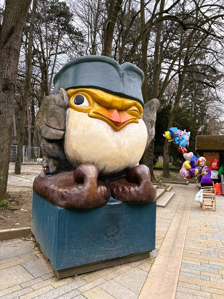 A large, whimsical statue of a bird wearing a hat, positioned in a park surrounded by trees. The bird has a cheeky expression and is perched on a pedestal. In the background, there are colourful balloons and other park elements.
