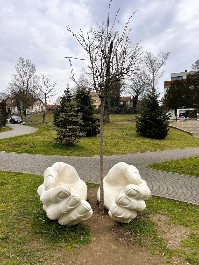A large sculpture of two white hands cupping a bare tree, set in a green park with walking paths and conifer trees in the background.